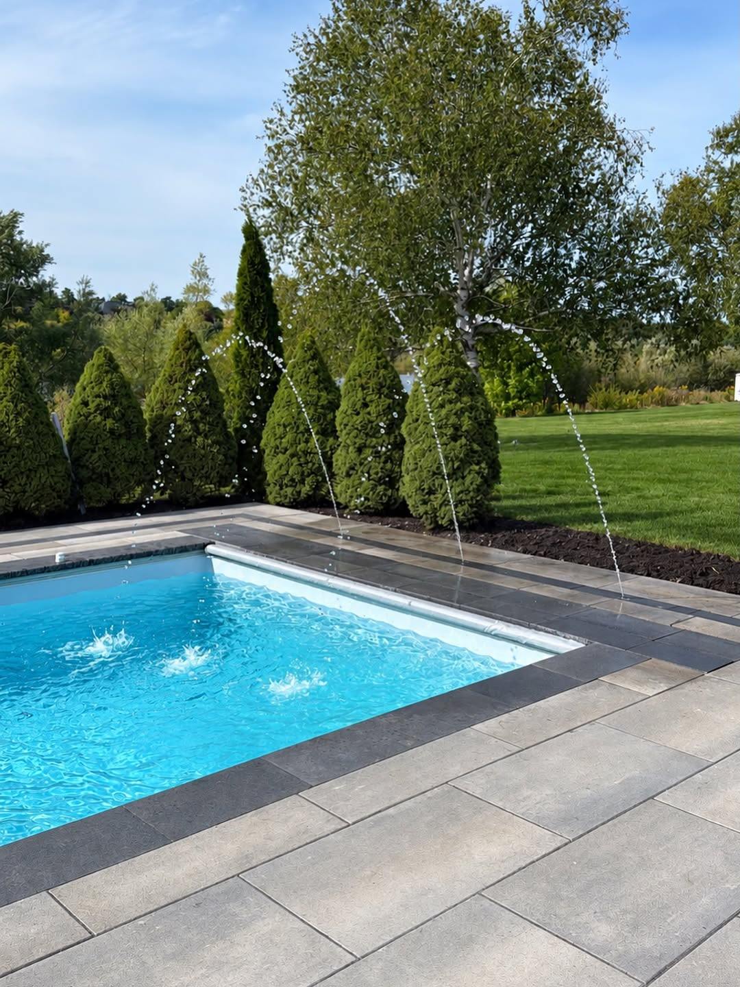 Fiberglass pool with deck jets beside a slab patio and cedars